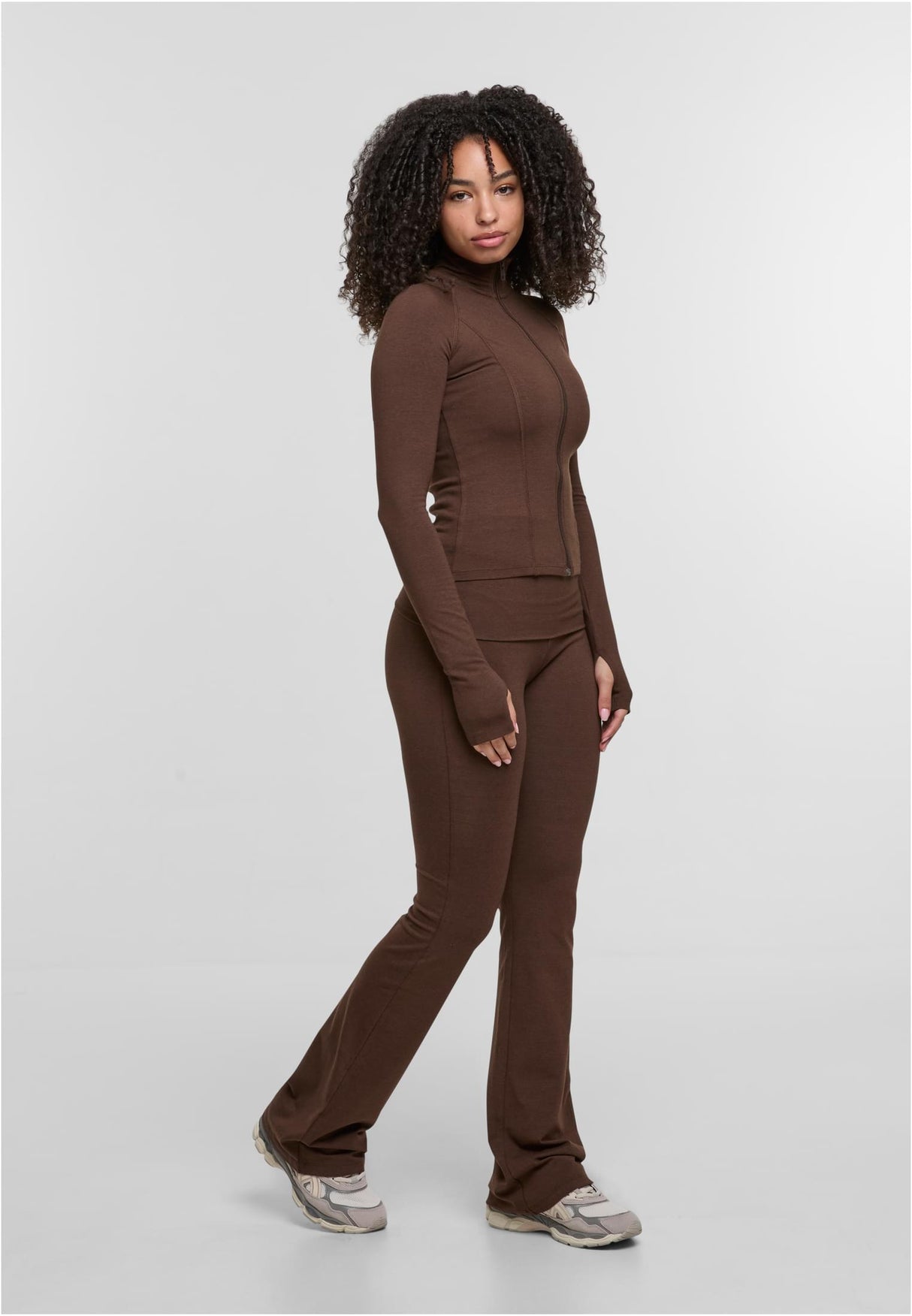 En kvinna poserar i en brun, figurnära träningsjacka med dragkedja och matchande utsvängda byxor.