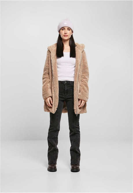 En kvinna som bär en öppen, beige UC Sherpa Jacket med svarta jeans, en vit topp och en ljusrosa mössa, sedd framifrån mot en ljusgrå bakgrund.